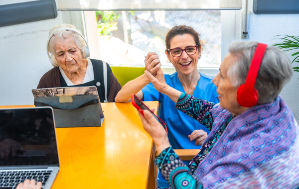 nursing assistant engaging with two elderly women in a care setting one woman using tablet while another enjoys music on headphones