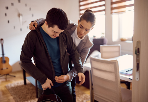 a caregiver assisting a young man in putting on a jacket in a bright room with a guitar in the background focused on support and care in everyday life