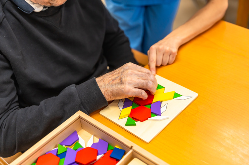 person helping elderly individual with colorful tangram puzzle on wooden tray two engaging minds